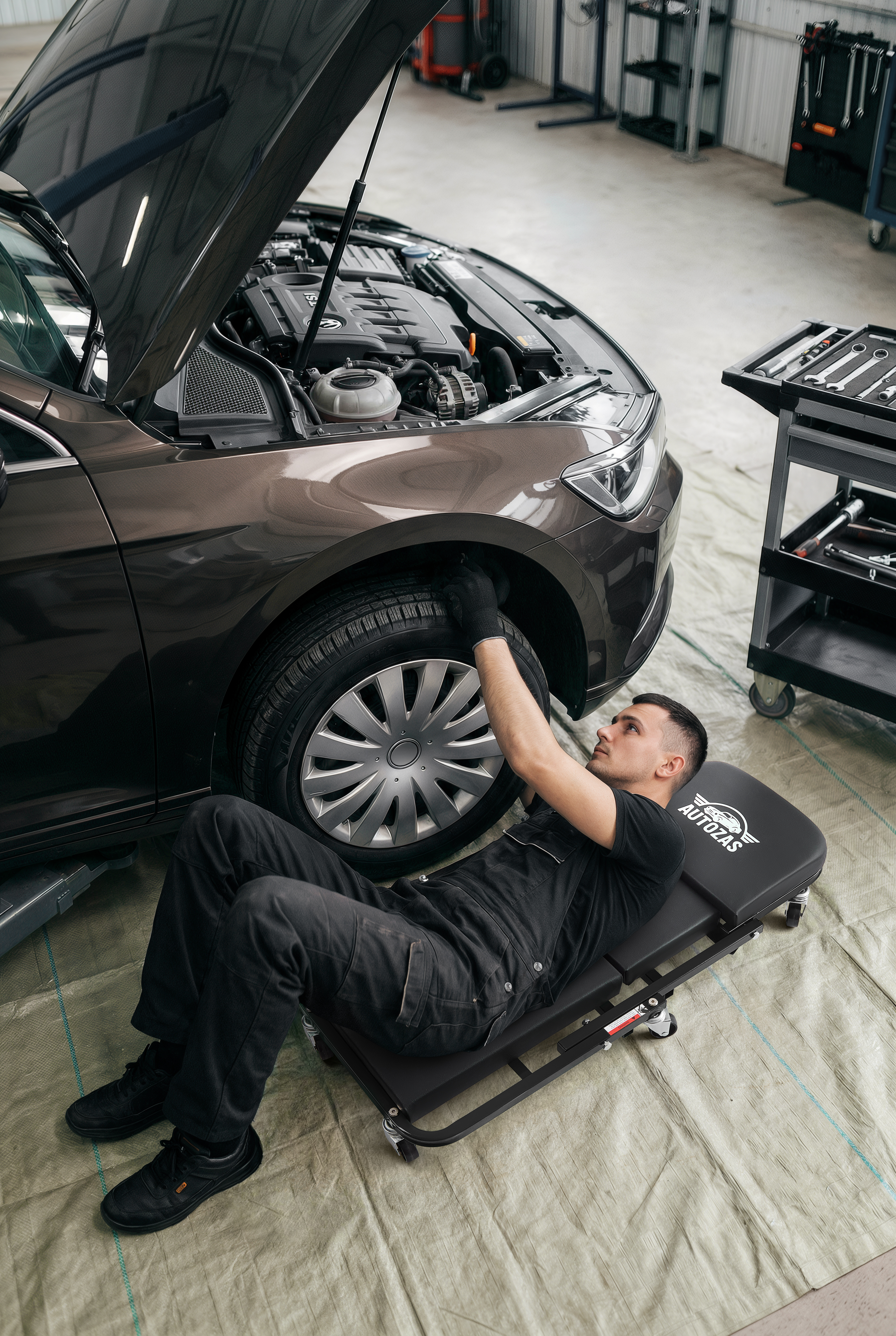 Professional mechanic lying on the Autozas Black creeper supporting full body weight — heavy-duty steel frame under real-world garage load